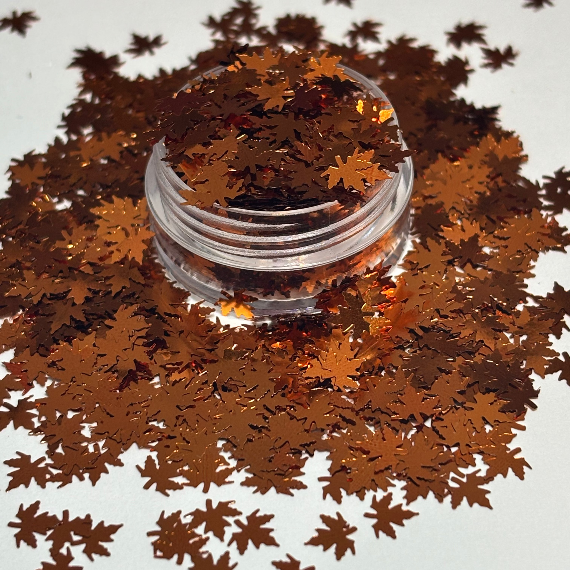 Shiny copper-brown metallic leaf-shaped confetti scattered around a small clear plastic jar on a plain white background.