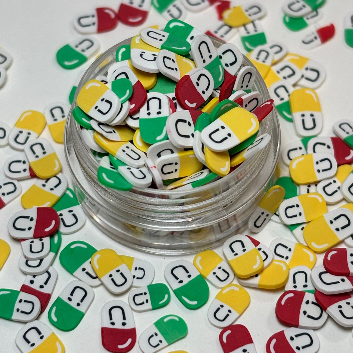 A clear round jar filled with small capsule-shaped charms or stickers in green, yellow, and red halves with white bottoms and black smiley faces, arranged on a white surface with more pieces scattered around.