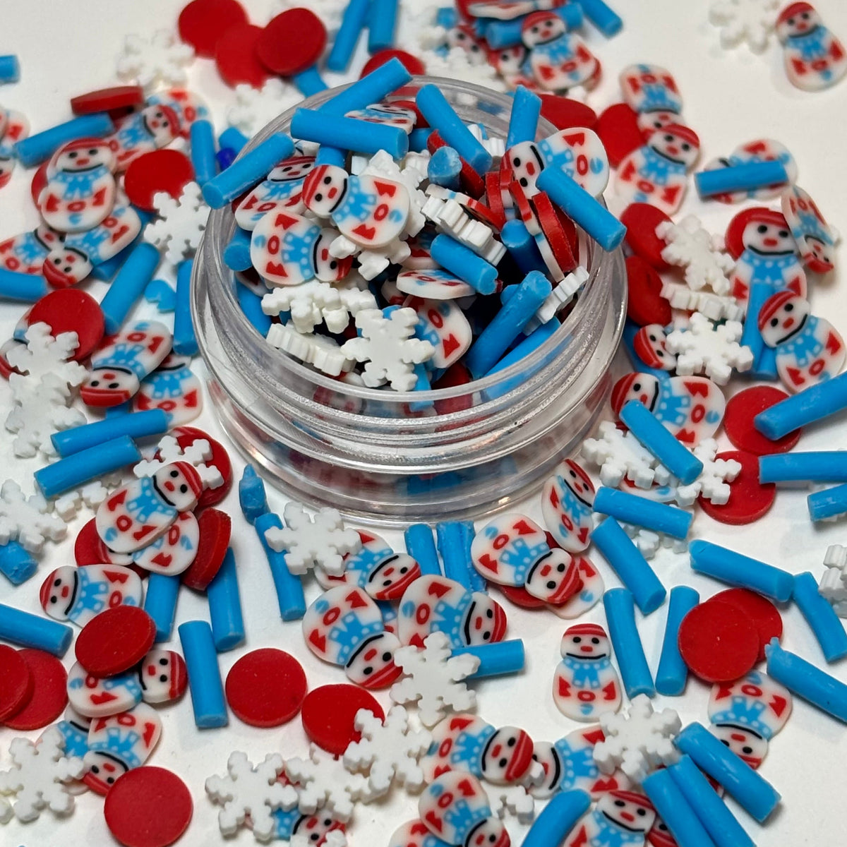 A clear round jar filled with small decorative sprinkles and surrounded by scattered red, white, and blue holiday-themed decorations on a white surface.