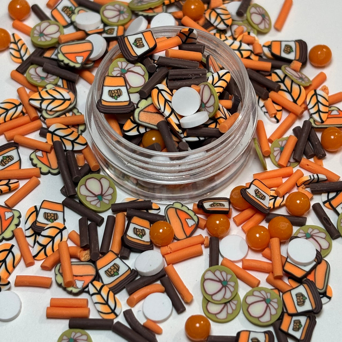 A clear round plastic jar filled with small decorative craft charms and sprinkles, surrounded by an abundance of matching orange, brown, white, and green decorative pieces on a white surface.