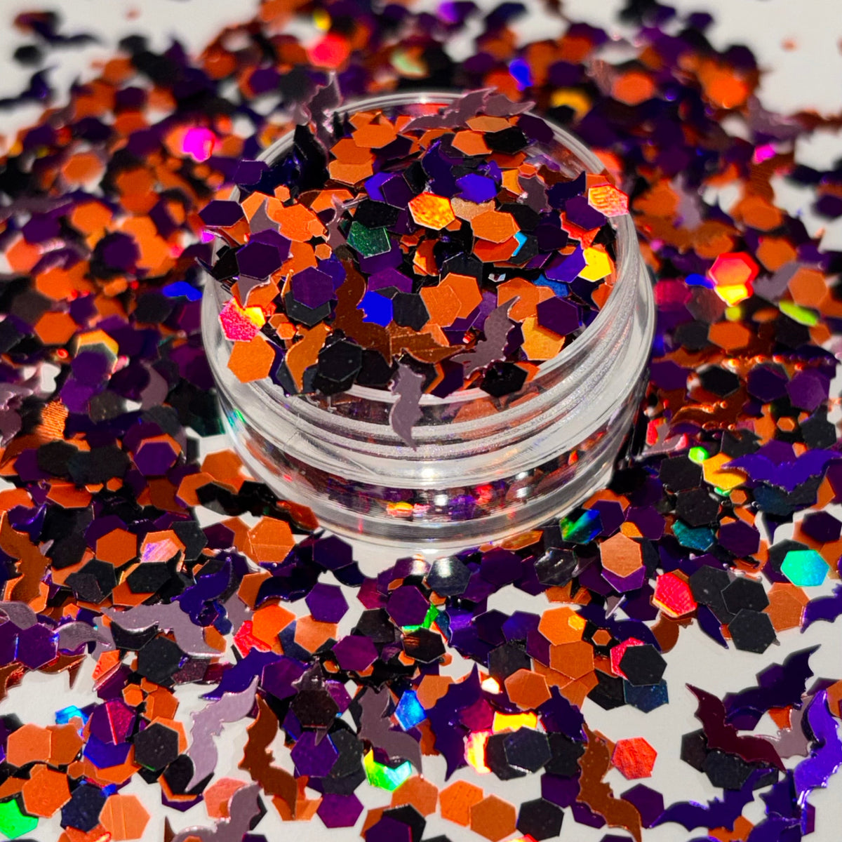 A small clear jar filled with iridescent hexagonal glitter, surrounded by spilled matching glitter on a white background.