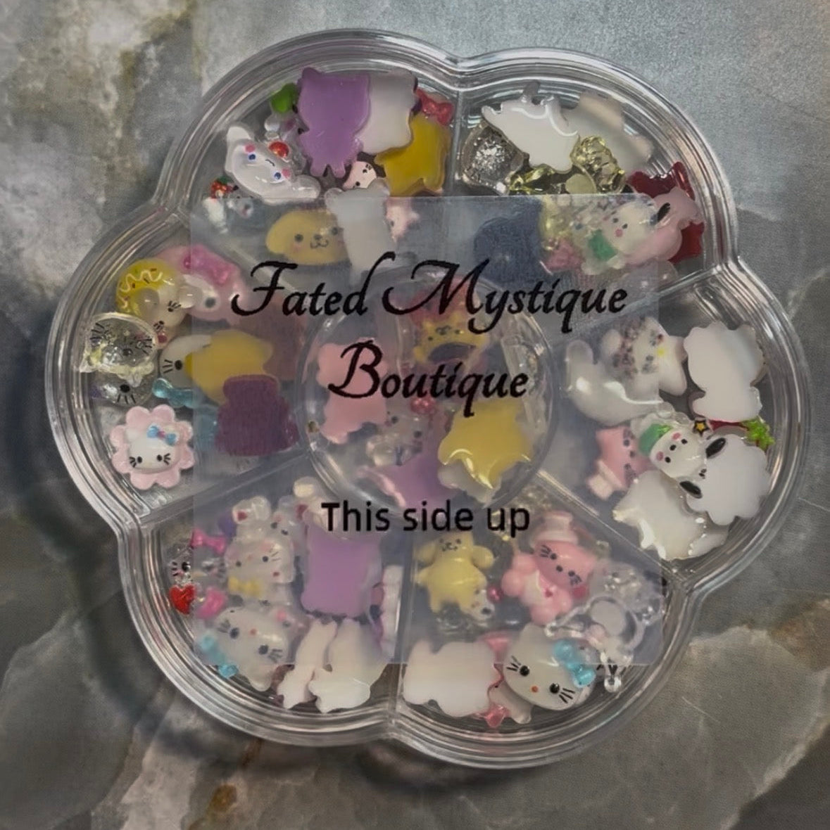 A flower-shaped clear plastic container filled with assorted small pastel charms, stickers, and embellishments, photographed on a gray marbled surface. The container has a translucent overlay reading 'Fated Mystique Boutique' and 'This side up'.